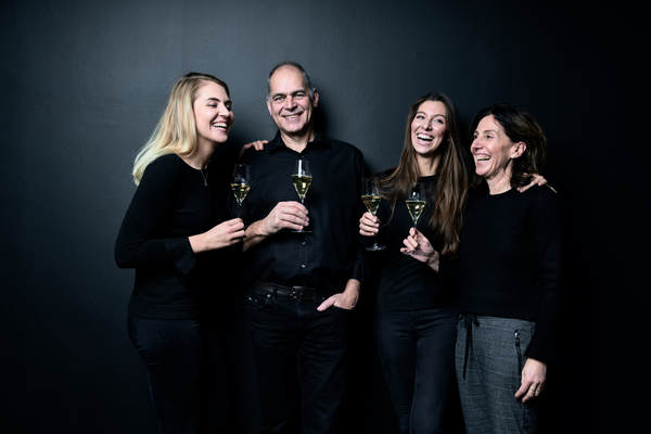 Vier Personen stehen vor einem schwarzen Hintergrund und halten Gläser mit Sekt. Sie lächeln und scheinen sich zu amüsieren, während sie miteinander sprechen oder lachen.