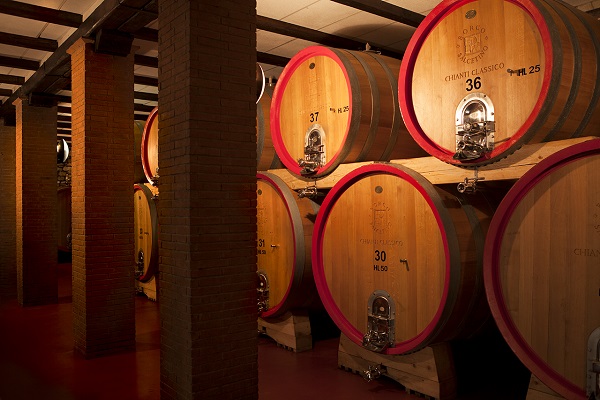 Große, runde Holzfässer stehen in einem Weinkeller. Die Fässer sind nummeriert und beschriftet, und zwischen ihnen sind massive Säulen aus Ziegelsteinen sichtbar, die den Raum unterteilen.