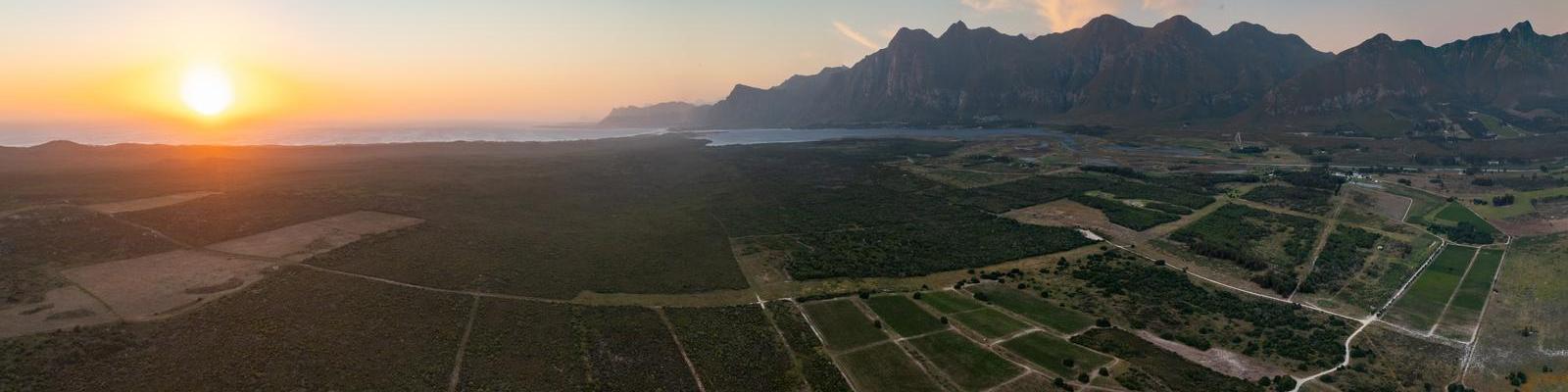 Eine Panoramaaufnahme zeigt einen Sonnenuntergang über einer weiten Landschaft mit Wiesen und Feldern. Im Hintergrund erkennt man Berge und die Küste des Ozeans.