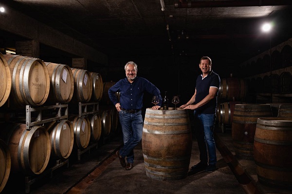 Zwei Männer stehen entspannt an einem Holzfass in einem Weinkeller. Im Hintergrund sind mehrere Holzfässer nebeneinander auf Regalen aufgestellt. Die Beleuchtung ist gedimmt.