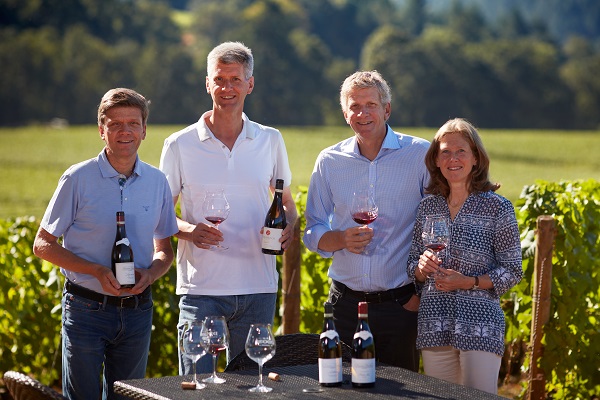 Vier Personen stehen im Freien in einem Weinberg. Sie halten Weinflaschen und Gläser mit Rotwein in der Hand, lächeln und posieren vor der malerischen Landschaft mit Weinreben im Hintergrund.