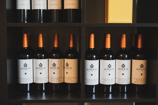 Auf einem Regal stehen mehrere Flaschen Wein mit orangefarbenen Kappen. Die Flaschen haben Etiketten mit dem Namen -Joaquin- und einem Stern. Oben befindet sich eine gelbe Box.