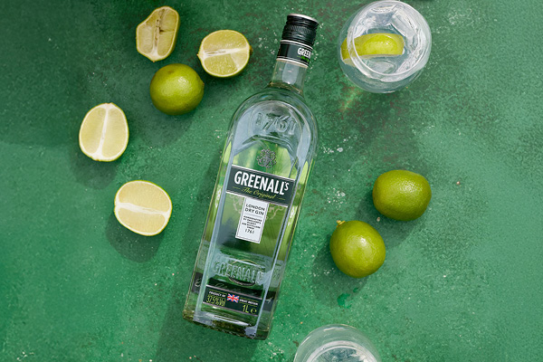 SDM_Juli_04_600 x 400 Eine Flasche Greenall-s Gin liegt auf einer grünen Oberfläche, umgeben von Limetten und Zitronenhälften sowie zwei Gläsern mit Wasser und Limettenscheiben.
