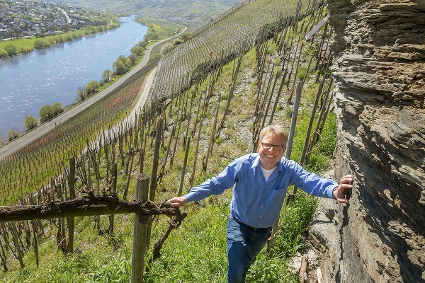 Ein Mann steht in einem Weinberg mit steilen Reihen von Reben. Hinter ihm fließt ein Fluss entlang der Ufer, und sanfte Hügel sind im Hintergrund sichtbar.