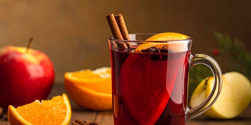 Zwei Gläser mit orangefarbenem Getränk stehen auf einem hellen Untergrund, dekoriert mit Sternanis und einer Zimtstange. Neben den Gläsern liegt eine halbe Orangenscheibe und ein Löffel.