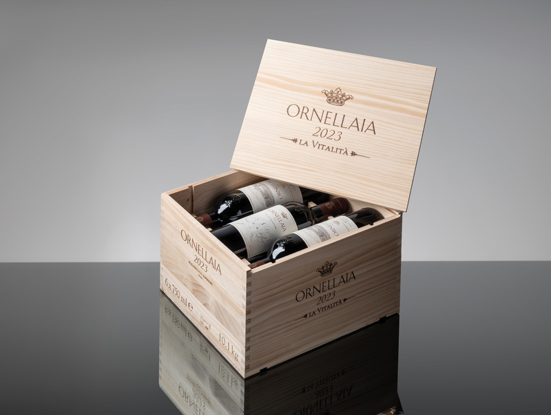 Eine offene Holzkiste mit dem Namen -ORNELLAIA 2023- enthält mehrere Weinflaschen. Die Kiste steht auf einer reflektierenden Oberfläche, die Umgebung ist neutral gehalten.