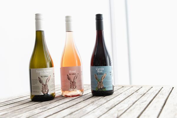 Drei Weinflaschen stehen auf einem Holztablett. Von links nach rechts: eine grüne Flasche mit Weißwein, eine rosafarbene Flasche mit Rosé und eine rote Flasche mit Rotwein, alle mit einem Design von einem Hasen.