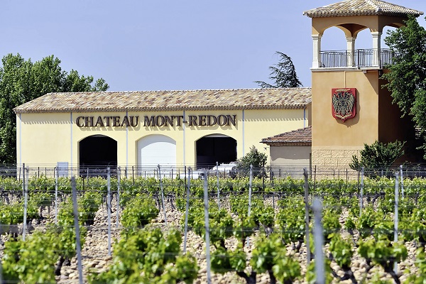 Mont-Redon Ein Weingut mit dem Schriftzug -Chateau Mont Redon- ist im Hintergrund zu sehen, umgeben von Weinreben. Ein Turm mit einem Balkon steht rechts, während Bäume im Hintergrund sichtbar sind.