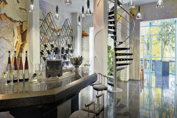 Ein modernes Bar-Innenraum mit einer eleganten Theke, Gläsern und Weinflaschen. Eine geschwungene Treppe führt nach oben und große Fenster bieten Blick ins Freie.