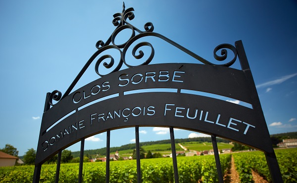 Ein schmiedeeisernes Tor mit der Aufschrift -CLOS SORBÉ DOMAINE FRANÇOIS FEUILLET- öffnet sich zu einem Weinberg. Im Hintergrund erstrecken sich grüne Felder unter einem klaren blauen Himmel.