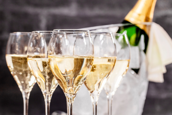 Fünf Gläser mit Schaumwein stehen in Reihe auf einem Tisch. Im Hintergrund ist eine Flasche Champagner in einem Eiseimer zu sehen, umgeben von einem weißen Tuch.