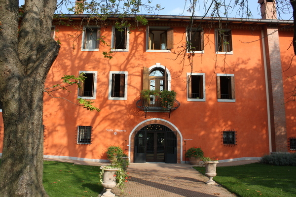 Ein orangefarbenes Gebäude mit mehreren Fenstern und einer großen, geschwungenen Eingangstür. Vor dem Haus stehen zwei Pflanzgefäße, umgeben von grünem Rasen und einem Baum im Vordergrund.