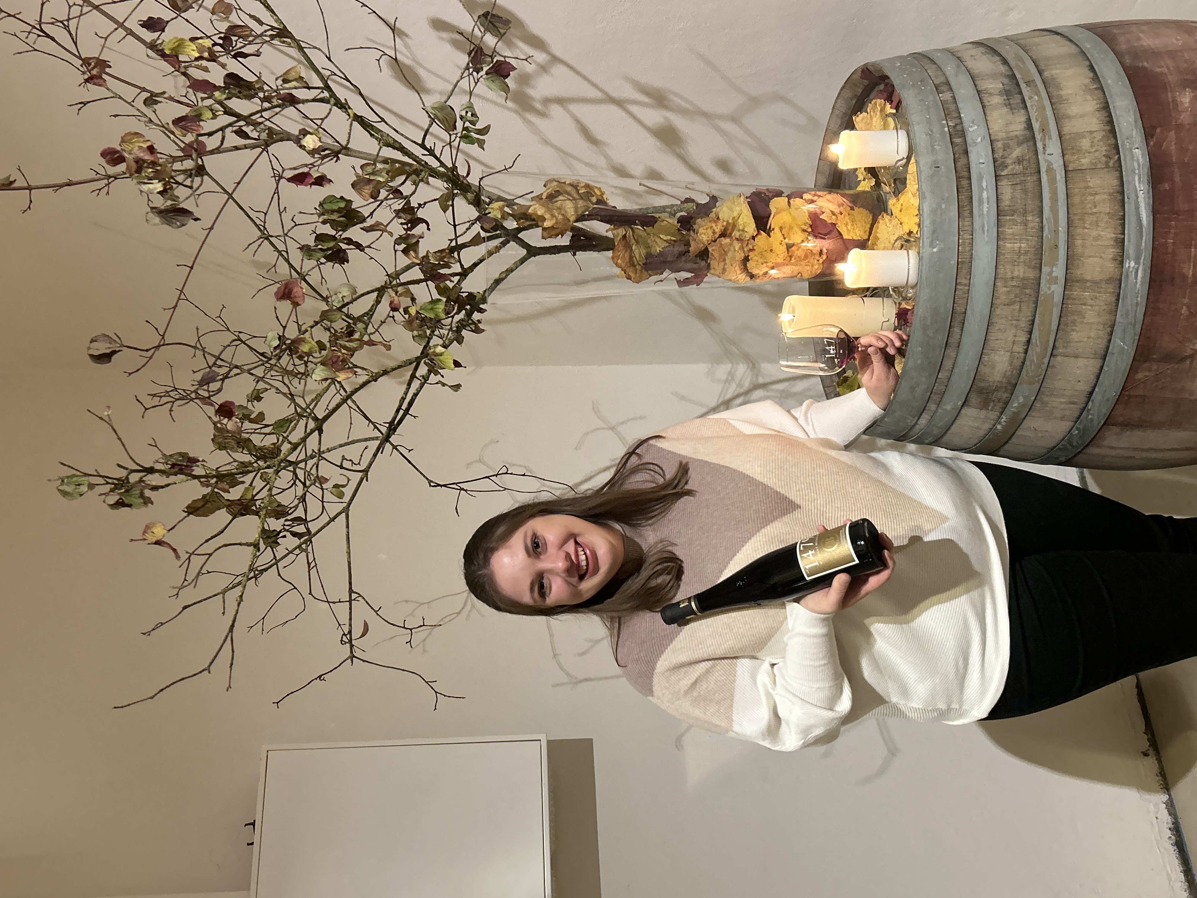 Eine Frau hält eine Flasche Wein in der einen Hand und ein Weinglas in der anderen, steht neben einem Holzfass, das mit Kerzen und Herbstblättern dekoriert ist.