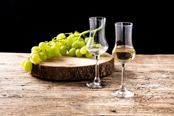 Ein Hand hält zwei Gläser. Das eine Glas enthält eine bernsteinfarbige Flüssigkeit, während das andere mit klarem Wasser gefüllt ist. Im Hintergrund sind unscharfe, helle Farben zu sehen.