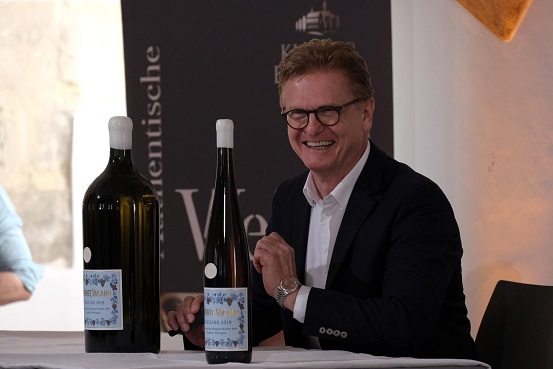Ein lächelnder Mann mit Brille sitzt an einem Tisch, vor ihm stehen zwei große Weinflaschen. Im Hintergrund ist ein Banner mit Aufschrift zu sehen.