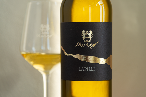 Eine Flasche Weißwein mit einem eleganten schwarzen Etikett und goldenen Akzenten steht im Vordergrund, während ein Weinglas mit goldenem Wein im Hintergrund unscharf zu sehen ist.
