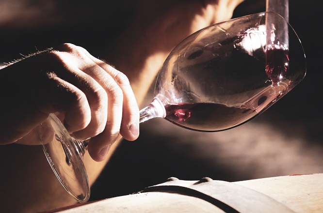 Ein Hand greift ein Weinglas, während ein roter Wein eingegossen wird. Im Hintergrund ist eine dunkle, holzverkleidete Umgebung zu sehen, die eine authentische Weintradition widerspiegelt.