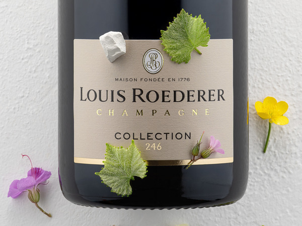 Eine Flasche Louis Roederer Champagner mit der Bezeichnung -Collection 246- liegt auf einem weißen Untergrund. Grüne Blätter und bunte essbare Blüten sind um die Flasche verteilt.