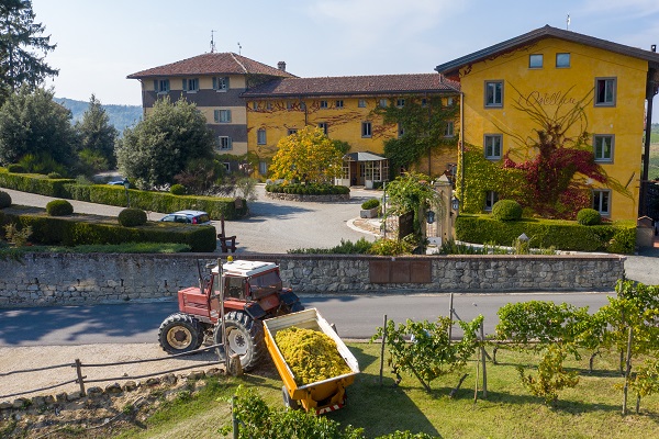 Ein roter Traktor entlädt eine Schaufel mit Trauben vor einem großen gelben Gebäude in einem ländlichen Gebiet. Rund um das Gebäude sind Pflanzen und ein gepflasterter Weg zu sehen.