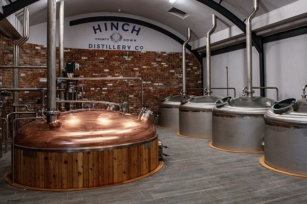 Kupferne und silberne Brennblasen stehen in einem modernen, industriellen Raum mit Ziegelwänden. Im Hintergrund ist der Schriftzug -HINCH COUNTY DOWN DISTILLERY CO- zu sehen.