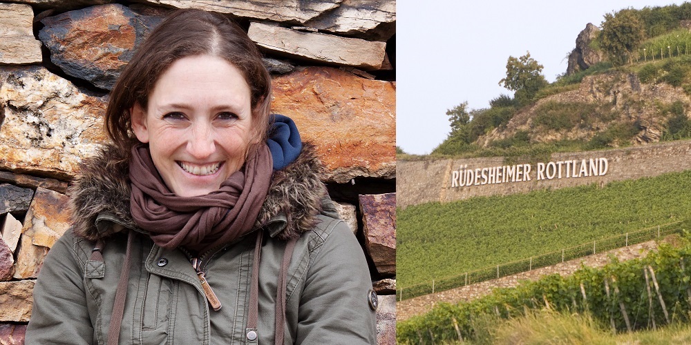 Eine lächelnde Person mit braunen Haaren und einem grünen Mantel steht vor einer Steinmauer. Im Hintergrund ist ein Weinfeld mit dem Schriftzug -Rüdesheimer Rottland- zu sehen.