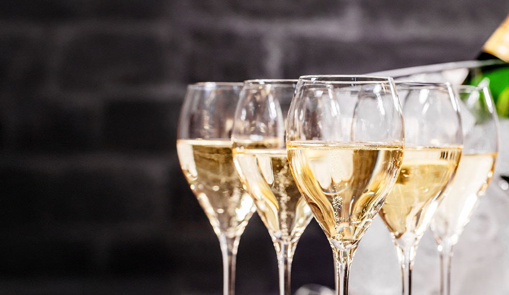 Fünf Gläser mit sprudelndem Sekt stehen in einer Reihe. Sie sind auf einem Tisch platziert, im Hintergrund ist eine dunkle Wand zu sehen.