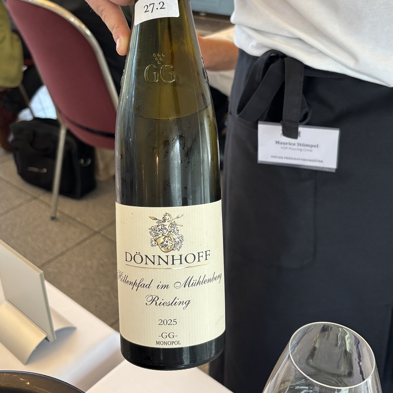 Eine Flasche Dönnhoff Riesling 2025 mit Etikett -Köllenpfad im Mühlenberg- wird von einer Person gehalten. Im Hintergrund sind Stühle und ein Tisch sichtbar.