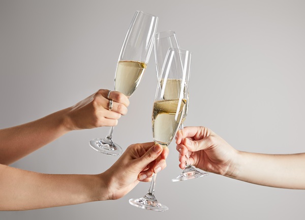 Sektgläser werden in einer Gruppe angestoßen. Die Hände der Personen sind sichtbar, während im Hintergrund das Wasser und ein Sonnenuntergang zu sehen sind.