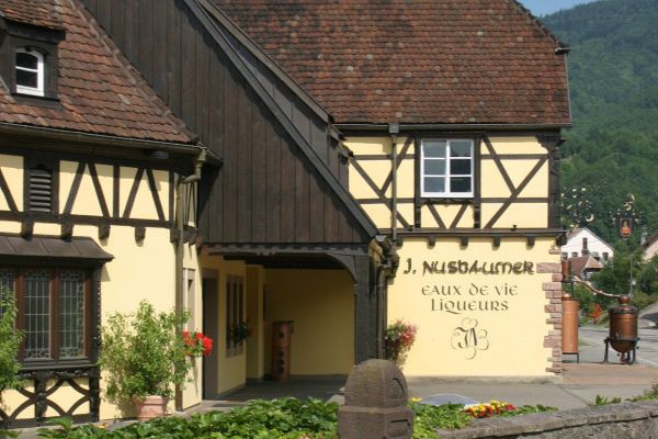 Ein Fachwerkhaus mit gelber Fassade und dunklem Holzoberteil. Vor dem Gebäude stehen Blumen und Pflanzen. An der Wand steht -J. Nusbaumer, eaux de vie, liqueurs-. Im Hintergrund sind Berge sichtbar.