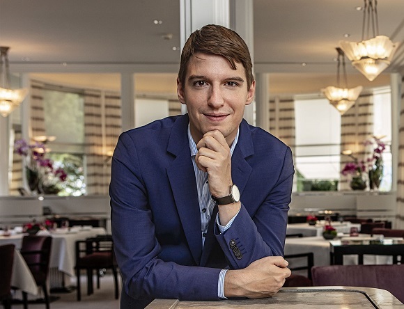 Ein junger Mann in einem blauen Anzug stützt sich auf einen Tisch in einem eleganten Restaurant. Der Raum ist geschmackvoll eingerichtet, mit Blumen und einem ansprechenden Lichtdesign.