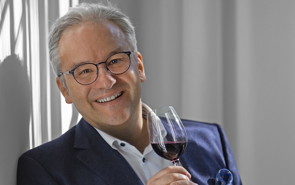 Auf einen Wein mit... Markus del Monego Ein lächelnder Mann hält ein Weinglas mit Rotwein in der Hand. Er trägt einen dunklen Anzug und eine helle Hemdbluse. Das Licht schafft interessante Schatten im Hintergrund.