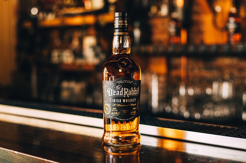 Eine Whiskeyflasche steht auf einer Bar. Die Flasche hat ein etiket mit dem Namen -Dead Rabbit Irish Whiskey-. Im Hintergrund ist eine unscharfe Barumgebung mit Gläsern zu sehen.