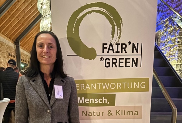 Eine Person steht neben einem Banner mit dem Text -FAIR-N GREEN- und den Worten -VERANTWORTUNG Mensch, Natur &amp; Klima-. Der Hintergrund zeigt eine rustikale Innenarchitektur mit einer Treppe.
