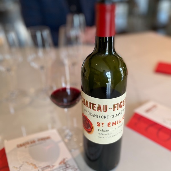 Eine Weinflasche mit dem Etikett -Château Figeac- steht auf einem Tisch, umgeben von Weingläsern und einigen roten Menükarten, während der Wein in einem Glas daneben sichtbar ist.