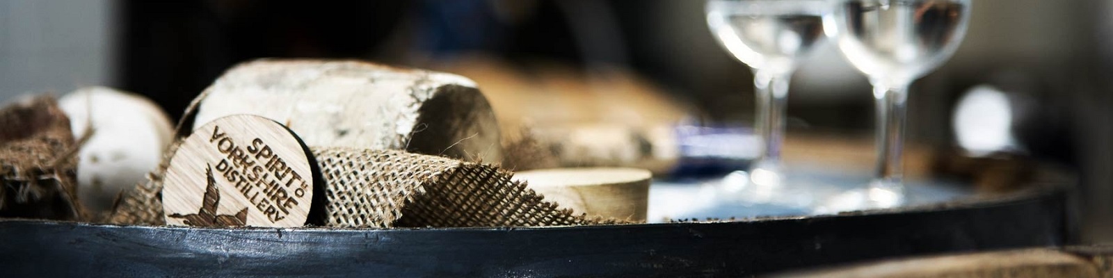 Spirit of Yorkshire Auf einem rustikalen Tisch liegen Holzstücke und eine Juteschnur. Im Hintergrund stehen zwei Weingläser mit klarem Inhalt. Ein Holzplättchen zeigt die Aufschrift -SPIRIT VORSPRUNG DISTILLERY-.