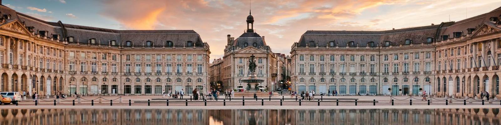 Ein beeindruckender Platz mit historischen Gebäuden ist zu sehen. Menschen schlendern um einen Brunnen, während sich bei Sonnenuntergang ein Farbenspiel am Himmel entfaltet. Der Platz spiegelt sich im Wasser.