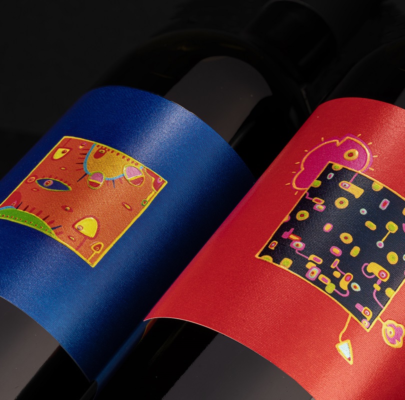 Zwei Weinflaschen stehen nebeneinander. Die linke Flasche hat ein blaues Etikett mit bunten, abstrakten Mustern, während die rechte Flasche ein rotes Etikett mit ähnlichen, lebhaften Designs trägt.