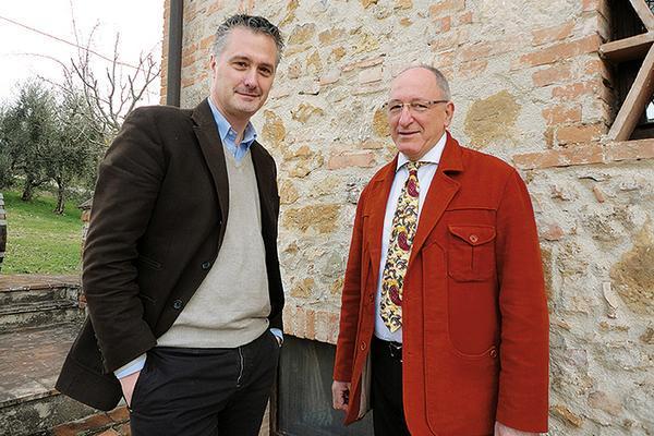 Zwei Männer stehen vor einer Mauer aus Stein und Ziegeln. Der jüngere Mann trägt einen dunklen Blazer und schaut zur Kamera, während der ältere Mann einen roten Mantel und eine bunte Krawatte trägt.