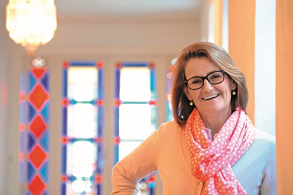Eine lächelnde Frau mit Brille trägt einen rosa Schal und steht in einem hellen Flur. Bunte Fenster mit geometrischen Mustern sind im Hintergrund zu sehen.