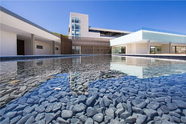 Ein modernes Gebäude mit einer glatten Wasserfläche im Vordergrund, die mit runden Steinen gefüllt ist. Der Himmel ist klar und blau, und die Architektur ist minimalistisch gestaltet.