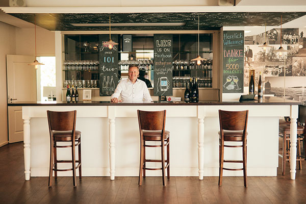Ein Kellner steht lächelnd hinter einer hellen Bar mit leeren Holzstühlen davor. Im Hintergrund sind Gläser und Flaschen zu sehen sowie mehrere Schiefertafeln mit Beschriftungen.