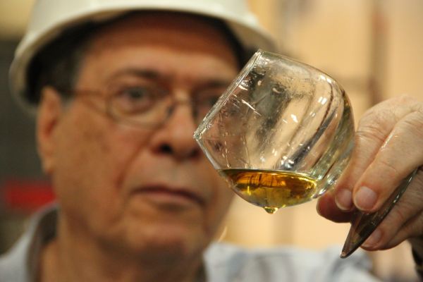 Ein älterer Mann mit Brille und einem weißen Bauhelm hält ein Glas mit goldfarbener Flüssigkeit. Er betrachtet die Flüssigkeit aufmerksam in einer industriellen Umgebung.