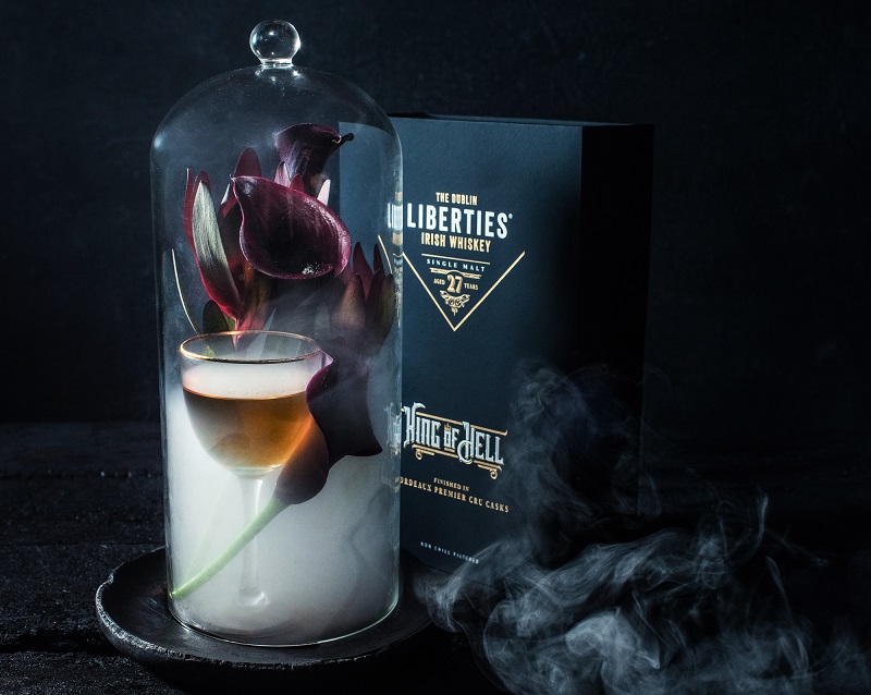 Ein Glas mit goldenem Getränk steht unter einer Glaskuppel. In der Kuppel befinden sich lila Blüten und Rauch. Im Hintergrund ist eine schwarze Whisky-Verpackung sichtbar.