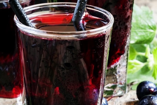 Transparente Gläser mit dunklem Getränk stehen nebeneinander, gefüllt mit einer schimmernden Flüssigkeit. Zwei Strohhalme stecken in einem Glas. Grüne Blätter und Beeren liegen im Vordergrund.