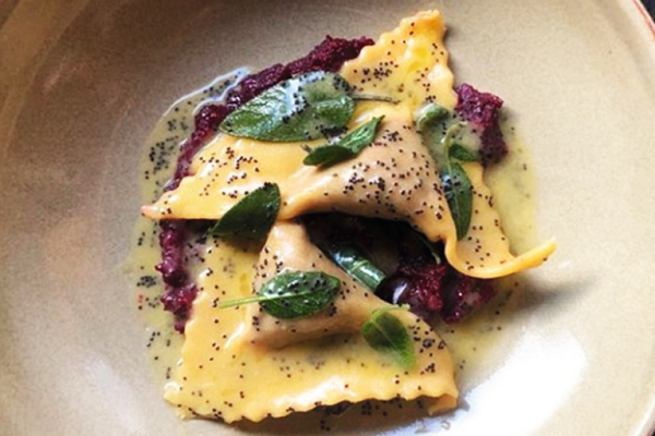 Hausgemachte Ravioli mit einer gelben Füllung liegen auf einem roten Püree, garniert mit Kräutern und Poppensamen. Die Sauce ist ölig und glänzend, auf einem großen Teller serviert.