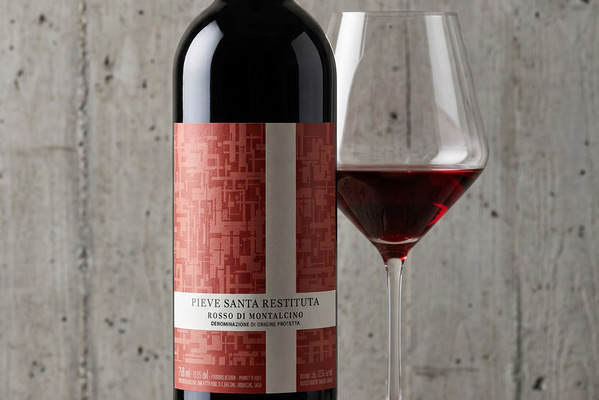 Eine elegante Weinflasche mit rotem Etikett steht neben einem Weinglas, das mit tiefrotem Wein gefüllt ist. Der Hintergrund ist eine helle, strukturierte Wand.