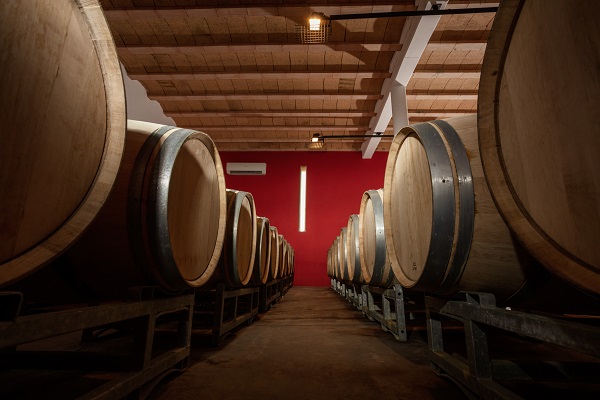 Fässer stehen in geraden Reihen auf Gestellen in einem Weinkeller. Durch das rote Wandpaneel und die Holzdecke entsteht eine warme, einladende Atmosphäre. Sanftes Licht beleuchtet den Raum.