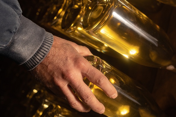 Roederer_Flaschen drehen_600x400 Ein Hand greift eine Glasflasche mit geronnener Flüssigkeit, umgeben von weiteren Flaschen in einer dunklen Umgebung, die schwaches Licht reflektiert.