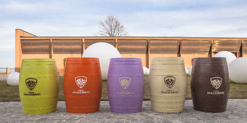 Fünf bunte Fässer stehen nebeneinander auf einem Steinboden. Jedes Fass ist in einer anderen Farbe gehalten und trägt das Logo -Pico Maccario-. Im Hintergrund sind große weiße Kugeln und ein Gebäude sichtbar.