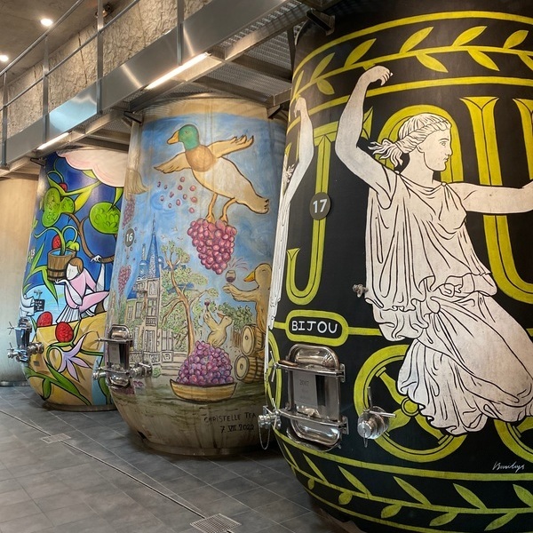 Drei große, bunt bemalte Weinfässer stehen in einem modernen Raum. Jedes Fass zeigt verschiedene Kunstwerke, darunter Tiere, Trauben und historische Figuren. Der Boden ist aus grauen Fliesen.